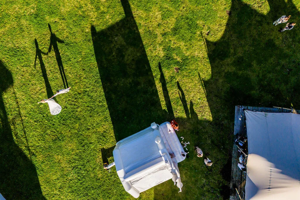 Dronefoto: de lange schaduwen van het bruidspaar op het gras naast de tent bij Zonnestraal