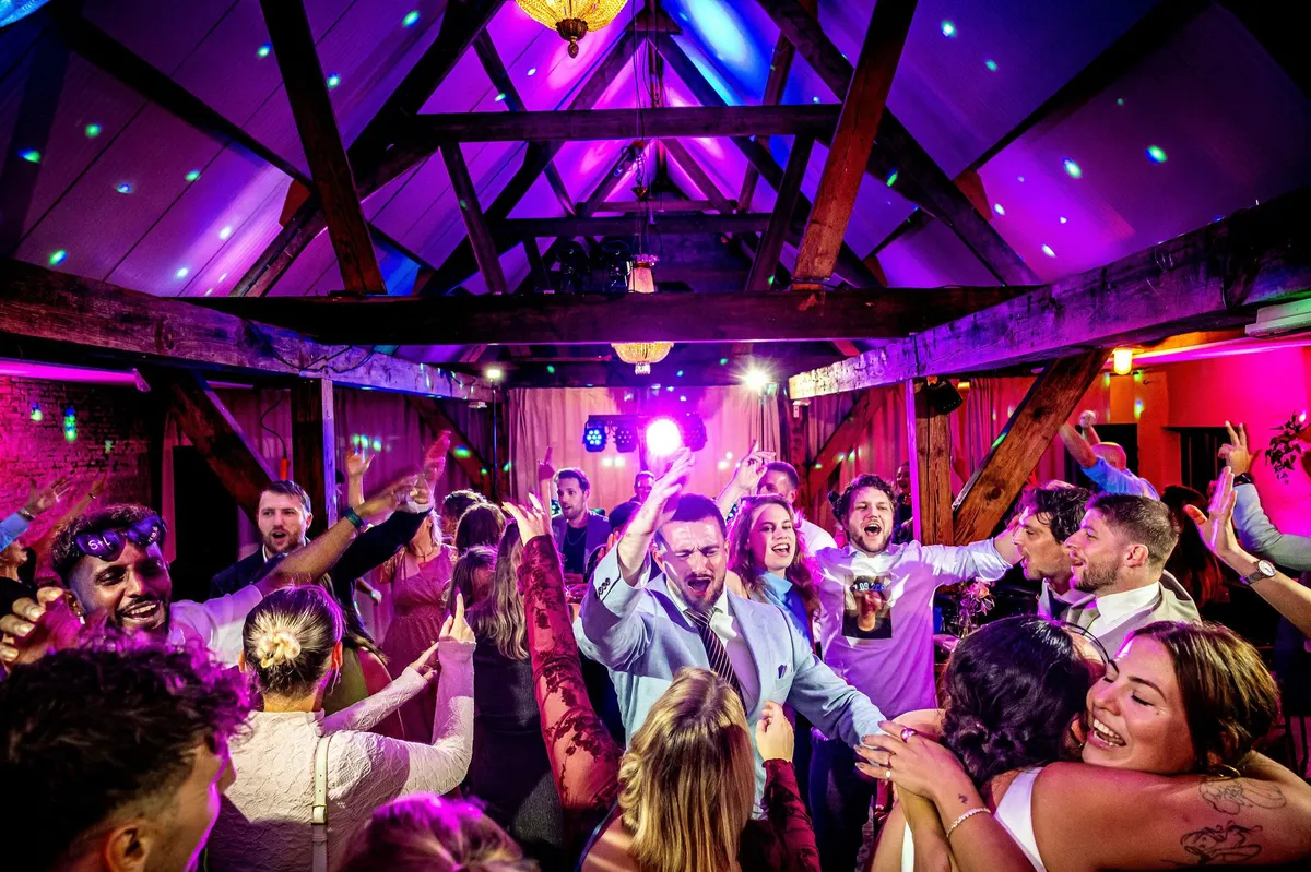 Een volle dansvloer onder houten balken en paars licht op het feest bij Le Farm
