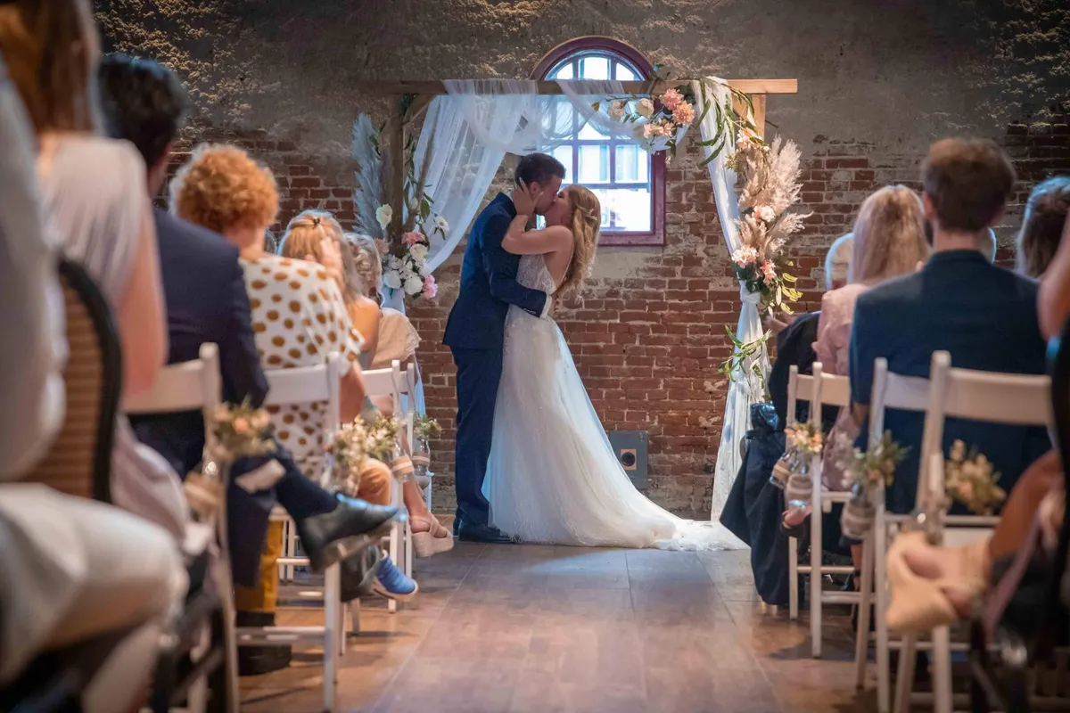 Het bruidspaar kust onder een bloemenboog in een bakstenen zaal met een boogvenster