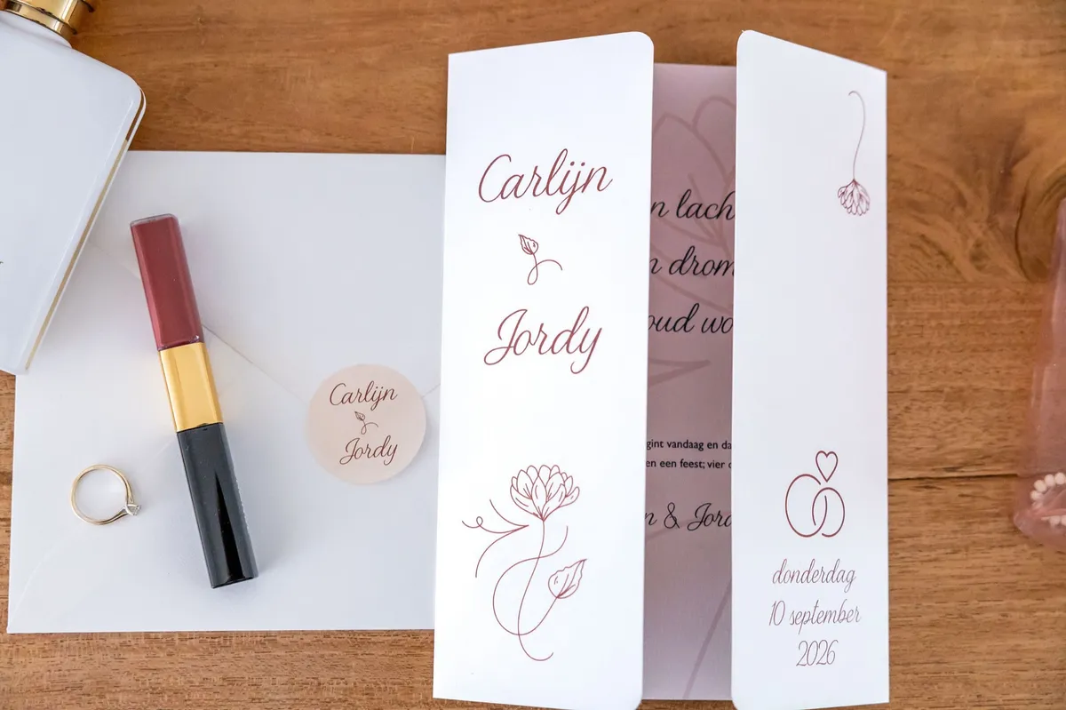Trouwkaart van Carlijn en Jordy met de trouwringen en een lippenstift op een houten tafel
