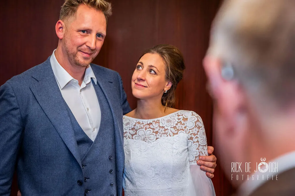 Het bruidspaar lacht naar een gast, de bruidegom met zijn arm om de bruid