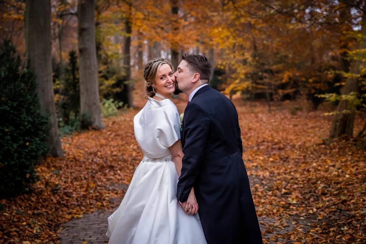 De bruidegom kust de bruid op haar wang in een bos vol herfstbladeren