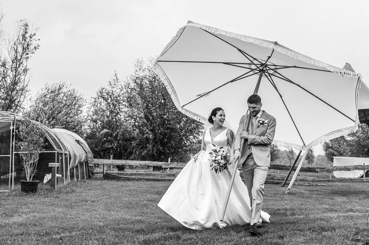 Het bruidspaar loopt lachend onder een grote parasol over het grasveld, langs de kassen (zwart-wit)