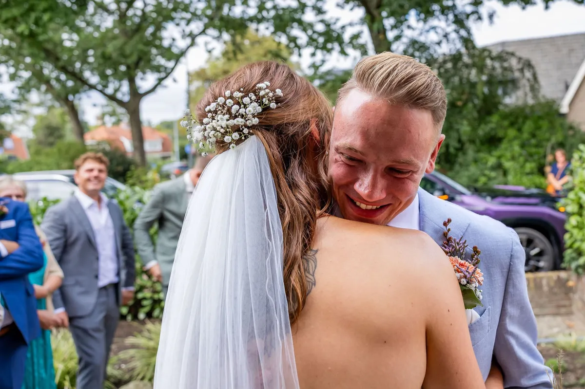 De bruidegom omhelst zijn bruid stralend bij het ophaalmoment, gasten kijken toe