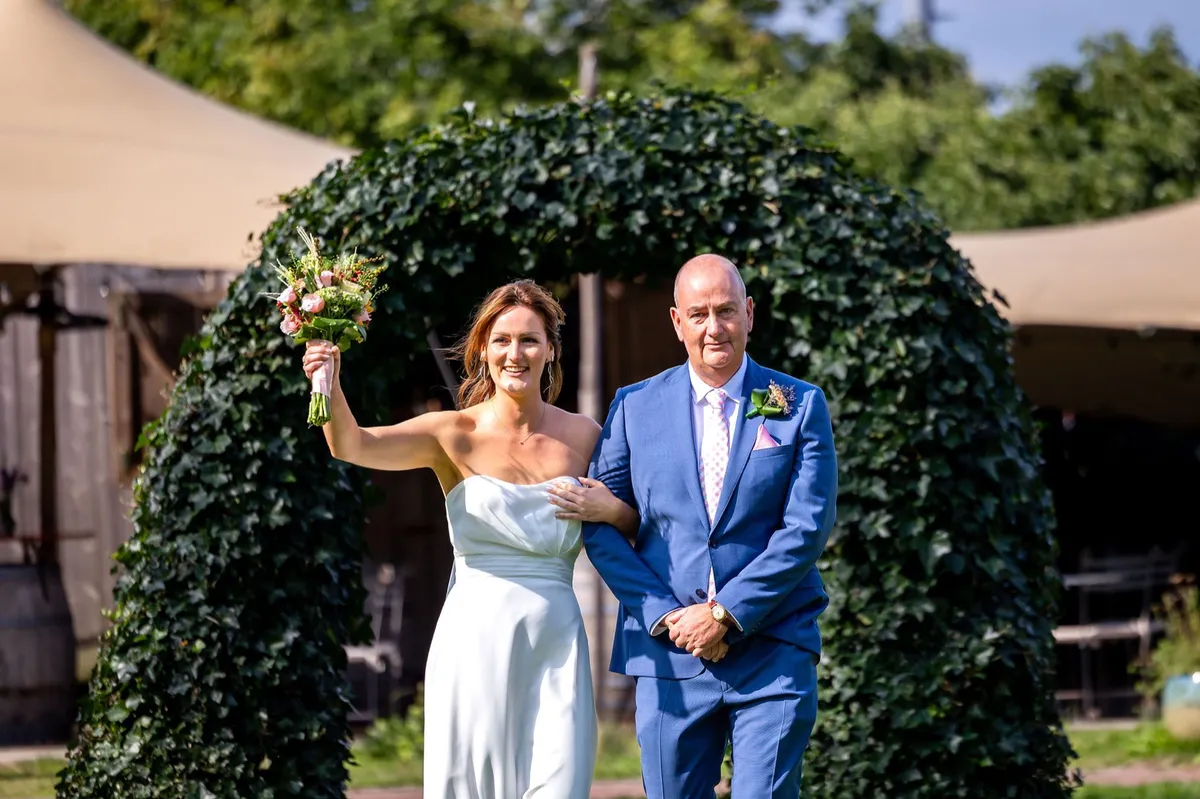 De bruid wordt door een groene boog van klimop naar de ceremonie bij Fier Bussum begeleid