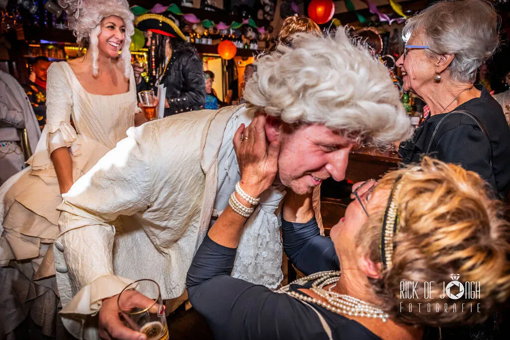 Een oudere vrouw knijpt een gast met een witte pruik in de wangen