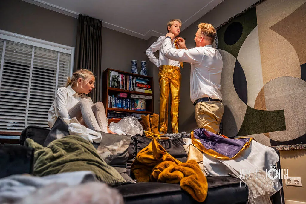 Een jongen in een gouden broek staat op de bank terwijl zijn kraag wordt rechtgetrokken