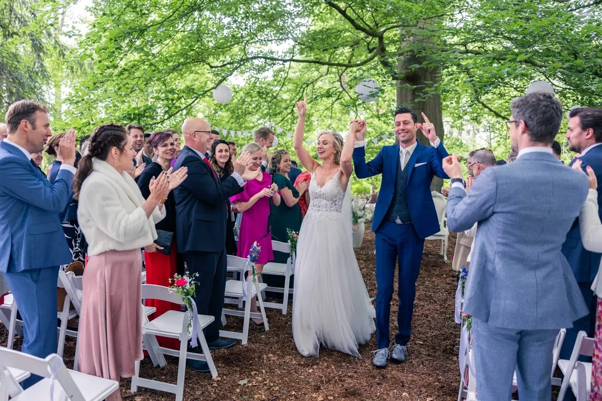 Het bruidspaar loopt juichend het gangpad af na een buitenceremonie onder de bomen