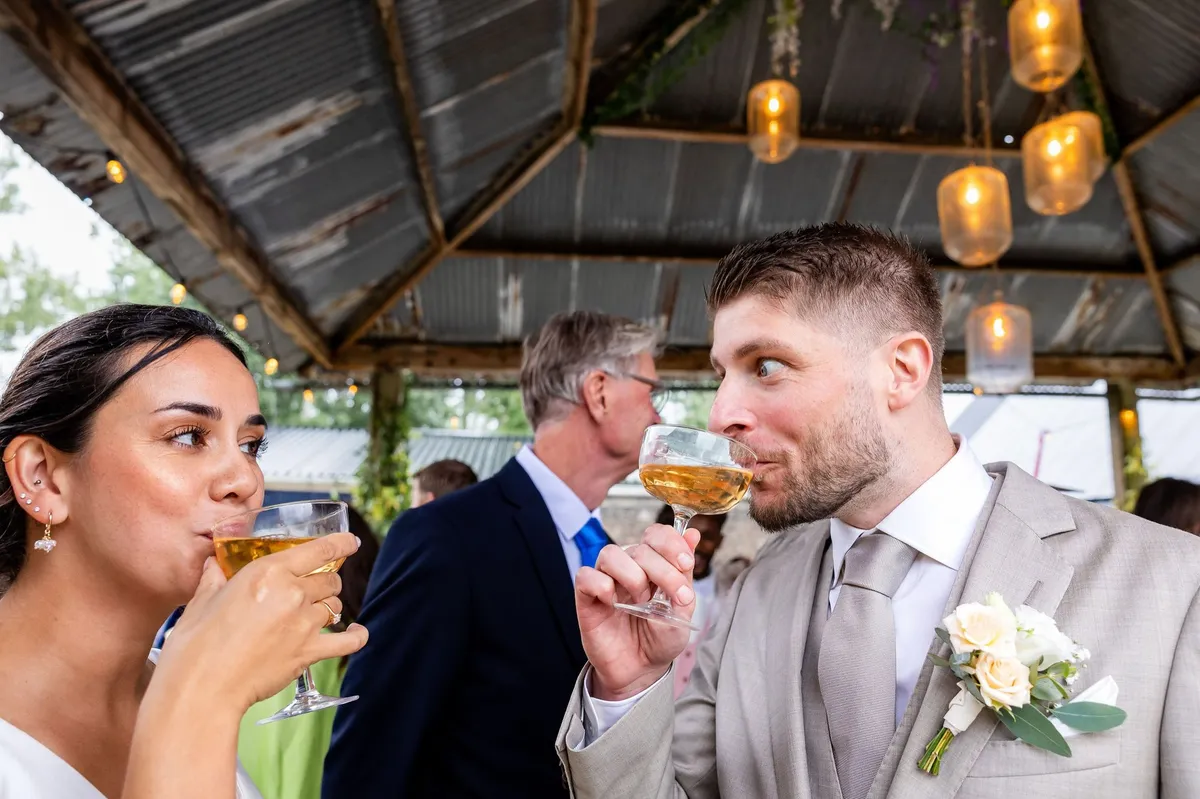 Het bruidspaar neemt een slok onder een overkapping met hanglampen bij Le Farm