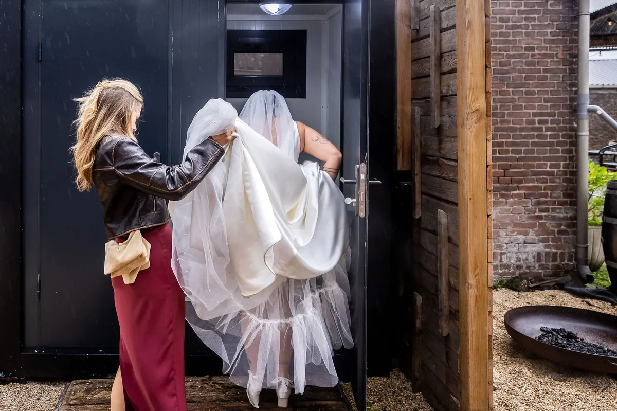De bruid tilt haar trouwjurk op en stapt door een deuropening van Le Farm, geholpen door een vrouw