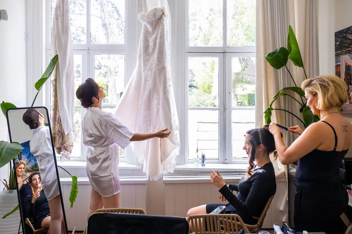 De bruid bekijkt haar trouwjurk die voor het raam hangt, terwijl een vriendin wordt opgemaakt