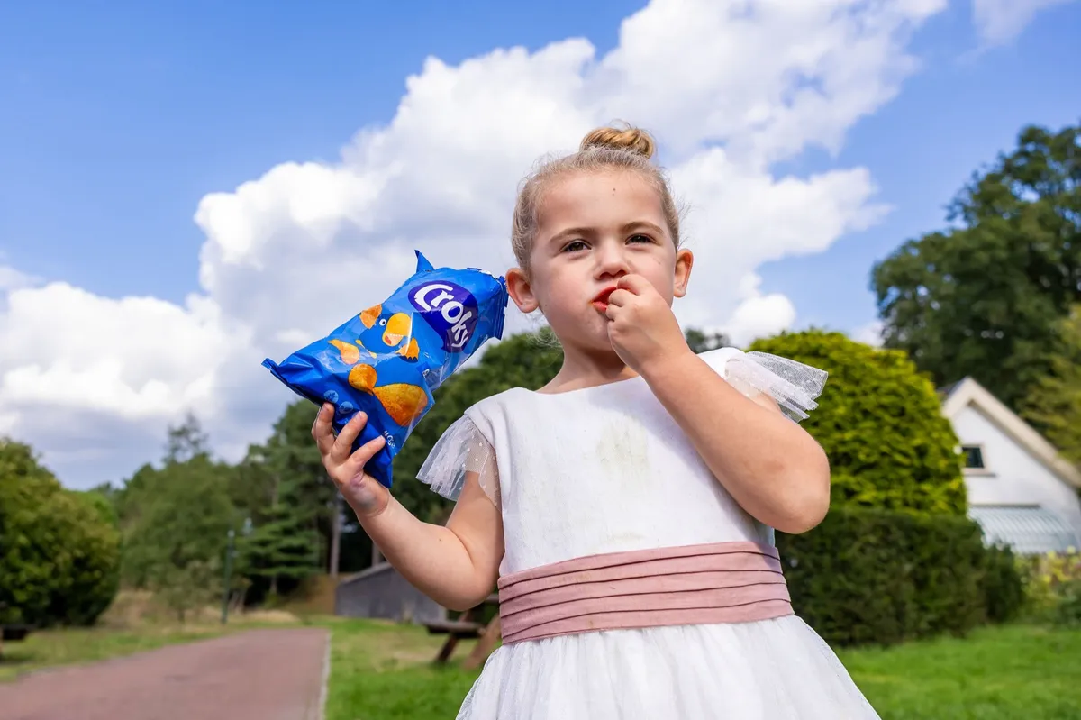 Bloemenmeisje in een wit jurkje met een zak chips op de trouwdag
