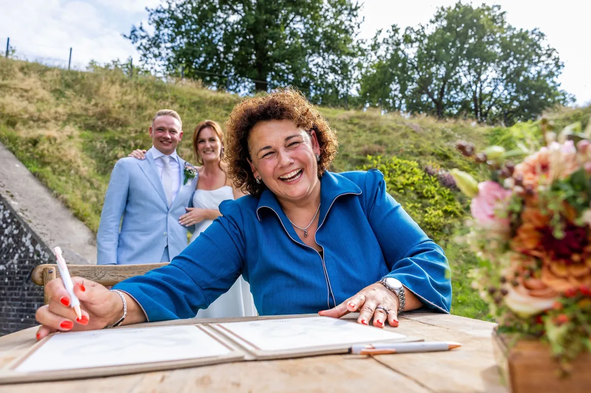 Babs Madelon zet lachend haar handtekening tijdens de trouwceremonie bij Fier Bussum, het bruidspaar kijkt toe